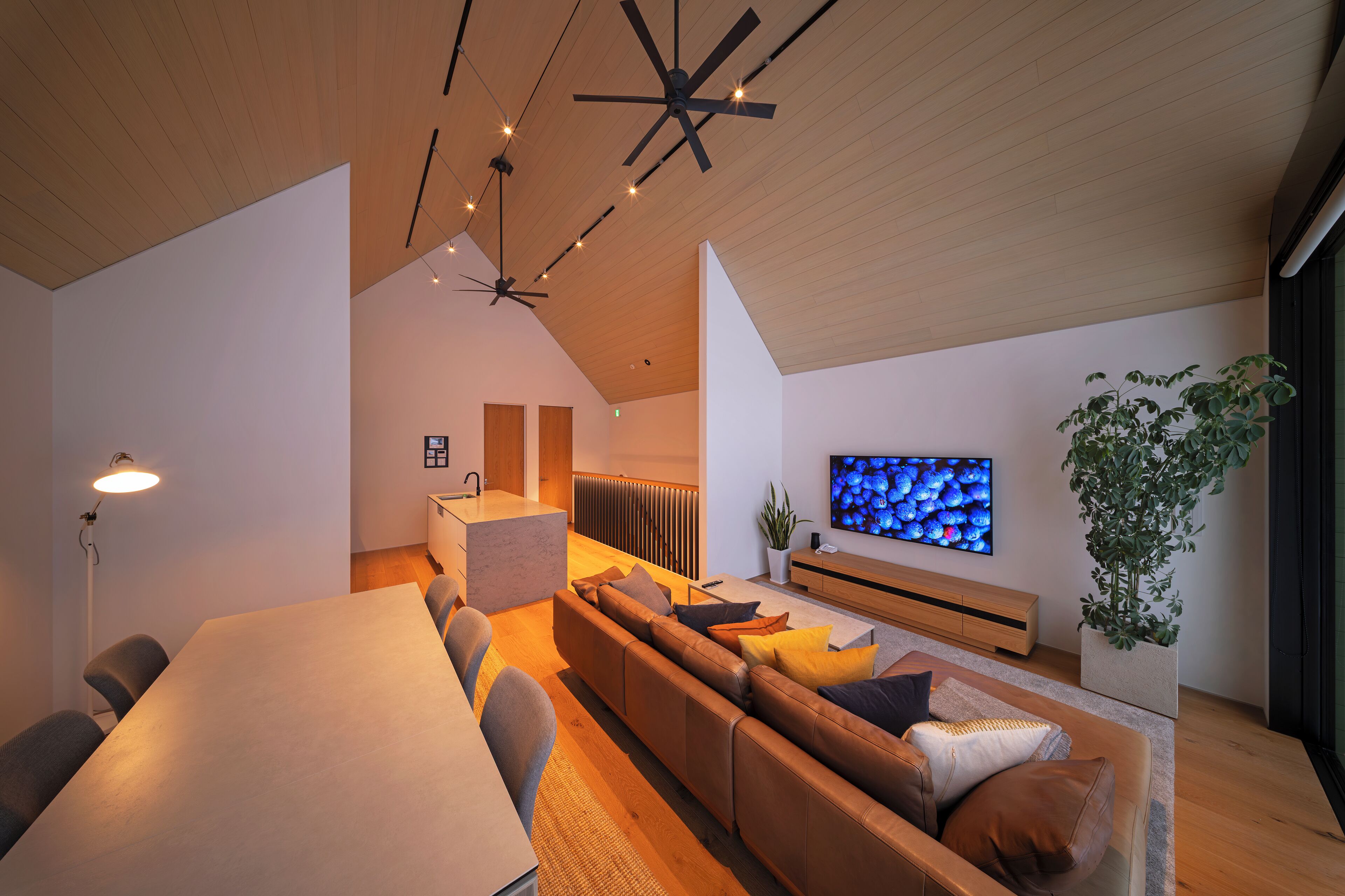 A modern living room with a large wooden dining table, a sofa with cushions, a television mounted on the wall, and a ceiling fan.