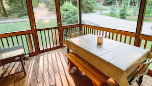 Varanda coberta com mesa e banco, com vista para uma rua residencial e vegetação.