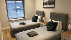 Ett sovrum med två sängar, ett fönster med persienner, ett element, ett nattduksbord med en lampa och en inramad tavla på väggen.