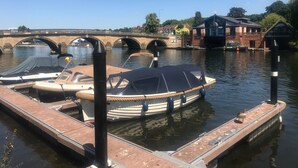 En kanal med båtar förtöjda vid en träbrygga, en bro i bakgrunden och byggnader längs flodstranden.