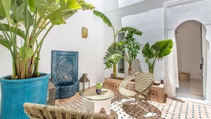 Eine sonnendurchflutete Terrasse mit einem großen Topfpflanzen, einem Korbstuhl, einem kleinen Tisch und einem mosaikgefliester Boden.