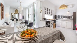 Ein modernes Wohnzimmer mit einer Couch, einem Esstisch mit Obstschale und einer Küchenzeile mit weißen Schränken und gefliestem Rücksprung.