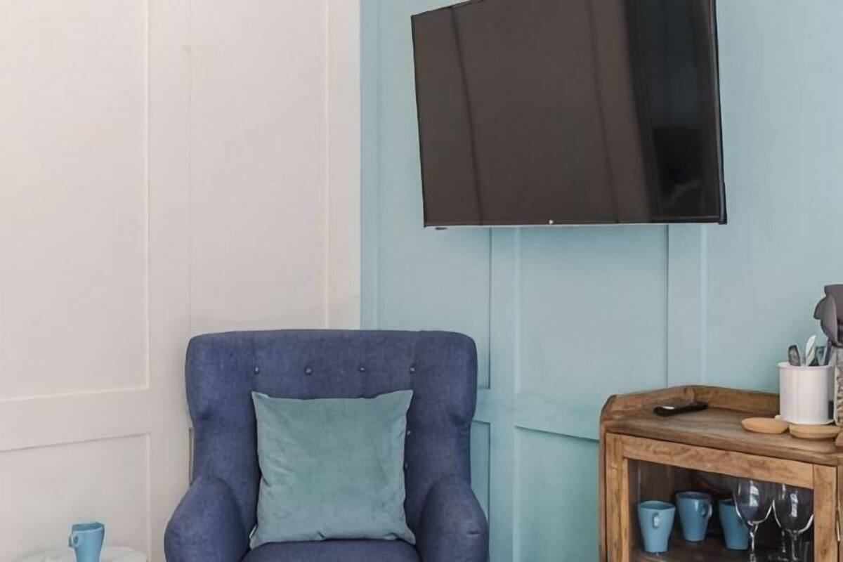 Ein Zimmer mit blauem Ohrensessel, einem Holznachttisch mit Tassen und Fernbedienung sowie einem an einer hellblauen Wand montierten Fernseher.