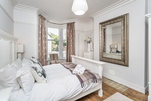 Une chambre à coucher bien rangée, avec un grand lit, une table de chevet, un miroir et une fenêtre avec des rideaux.