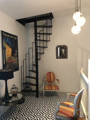 Un escalier en colimaçon, une chaise, une table avec une lampe, un tableau encadré et un sol à motifs.