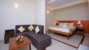 A hotel room with a sofa, a bed, a wooden nightstand, and a small table with a flower arrangement.