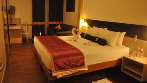 A hotel room with a bed, a bedside table with a lamp, a chair, a desk with a computer, and a TV mounted on the wall.