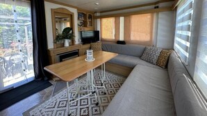 A compact living space with a sofa, a small table, a television, and a window with blinds. - Gîte Mobil-home in Drôme Provençale (Saint-Pantaléon-les-Vignes)