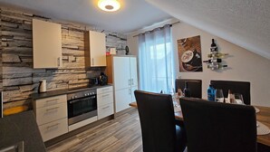 A modern kitchen with a stone wall backsplash, white cabinets, and a dining area with a table and chairs. - Fe-Wo Gold-Blick*** (Korbach)