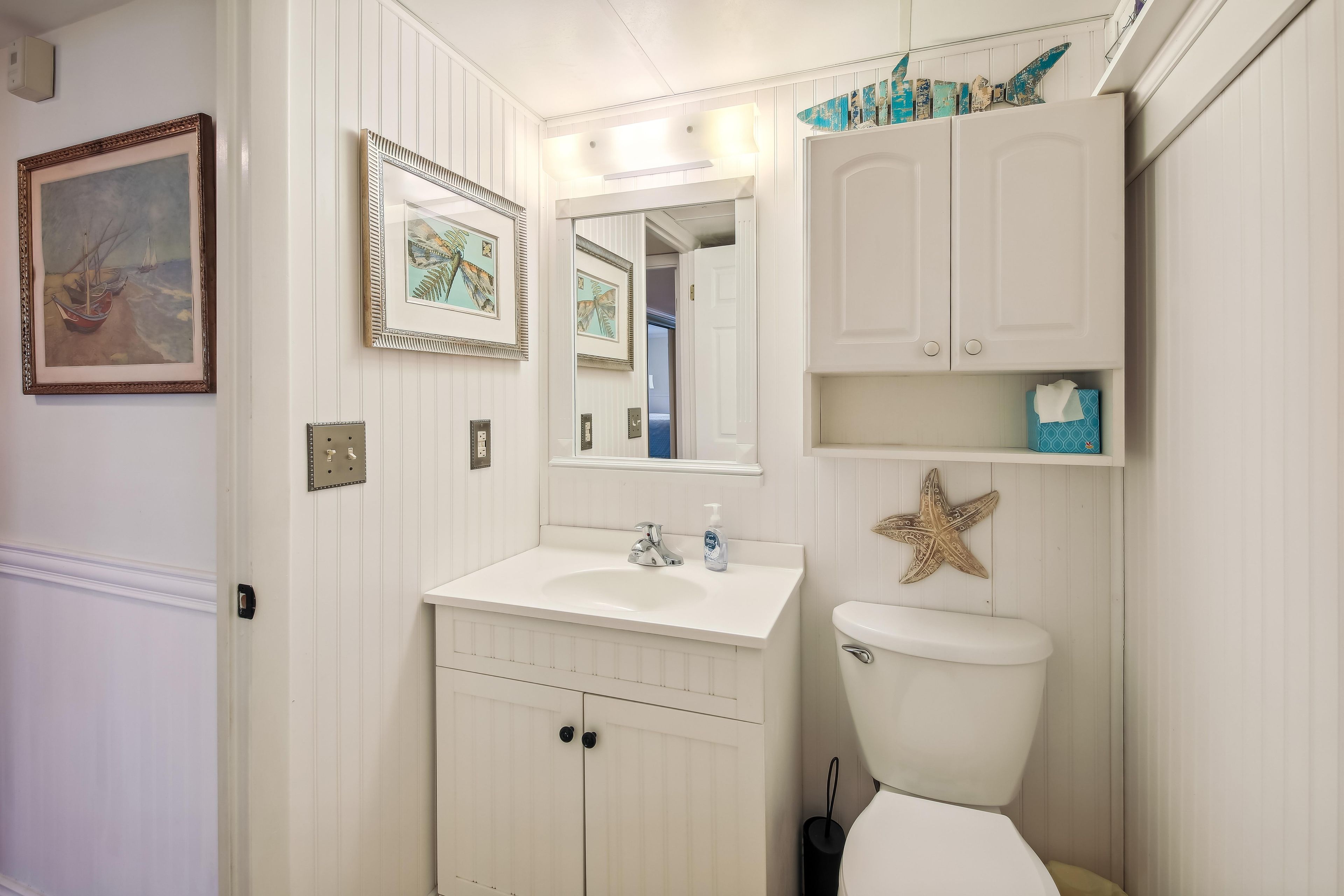A bathroom with a white sink, mirror, and toilet. There are framed pictures on the wall and a decorative starfish on the wall.