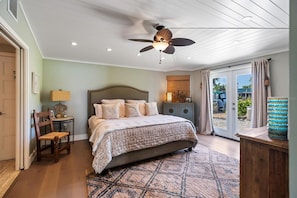 Ein Schlafzimmer mit Bett, Deckenventilator, Nachttisch, Stuhl, Lampe, Schrank und Blick ins Freie durch eine Schiebetür.