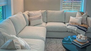 A modern living room with a sectional sofa, a neon sign, a window with blinds, and a coffee table with a bowl of cookies.
