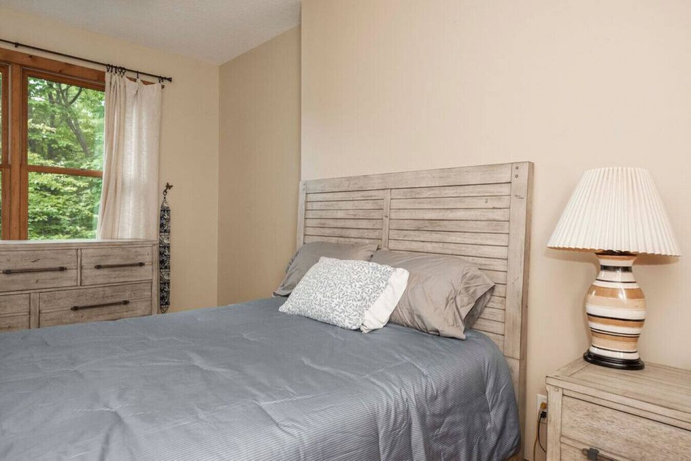 A bedroom with a bed, a nightstand, a lamp, a window with curtains, and a dresser.