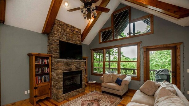 A cozy living room with a stone fireplace, wooden bookshelves, a beige sofa, and large windows offering a view of the outdoors. - Nature lovers paradise in Connestee Falls 14 minutes to Brevard! (Brevard)