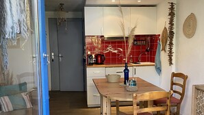 A dining area with a wooden table and chairs, a kitchen in the background with red tiles, and a door leading to another room.