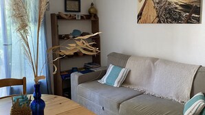 A cozy living room with a wooden table, a grey sofa, a vase with dried plants, and a framed picture on the wall.