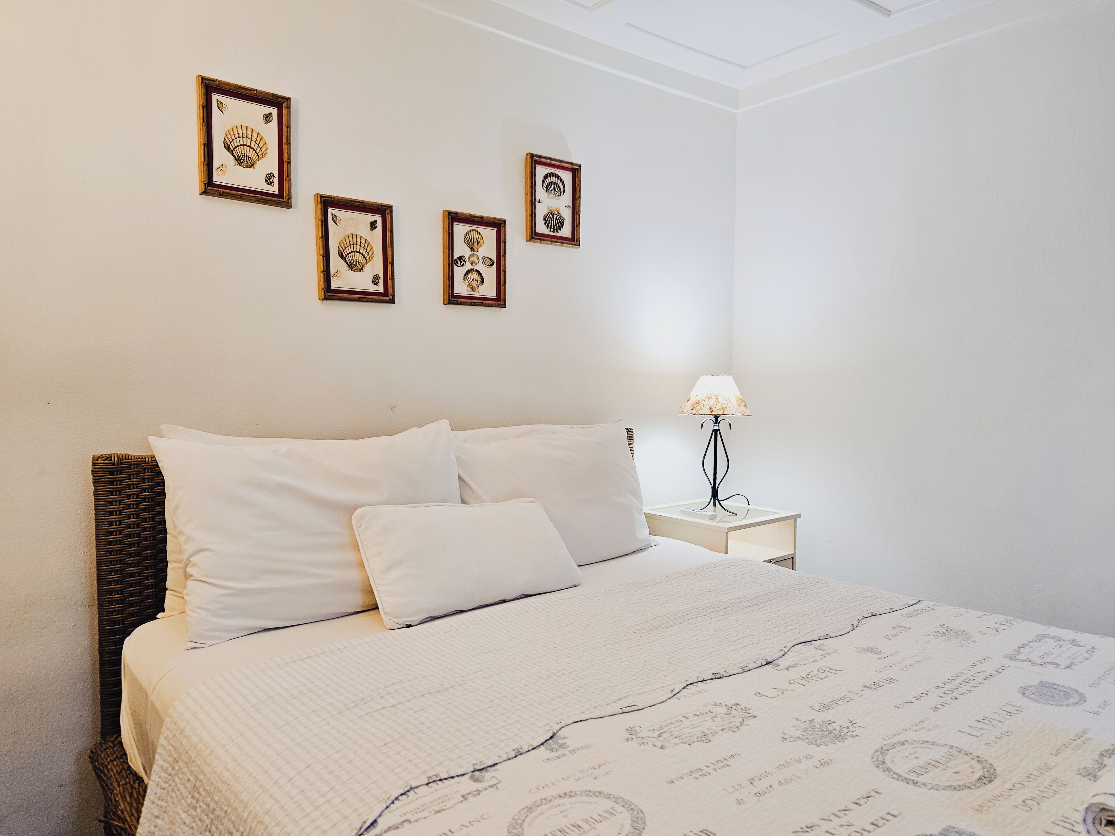 Un lit bien fait, avec du linge de lit blanc et une couvre-lit à motifs, une table de chevet avec une lampe, et trois tableaux encadrés accrochés au mur.