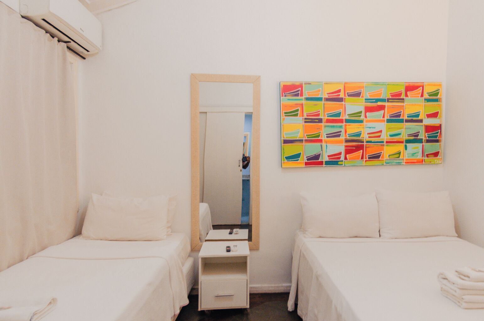 Habitación de hotel con dos camas individuales, una mesita de noche con lámpara, un espejo y una obra de arte colorida en la pared.