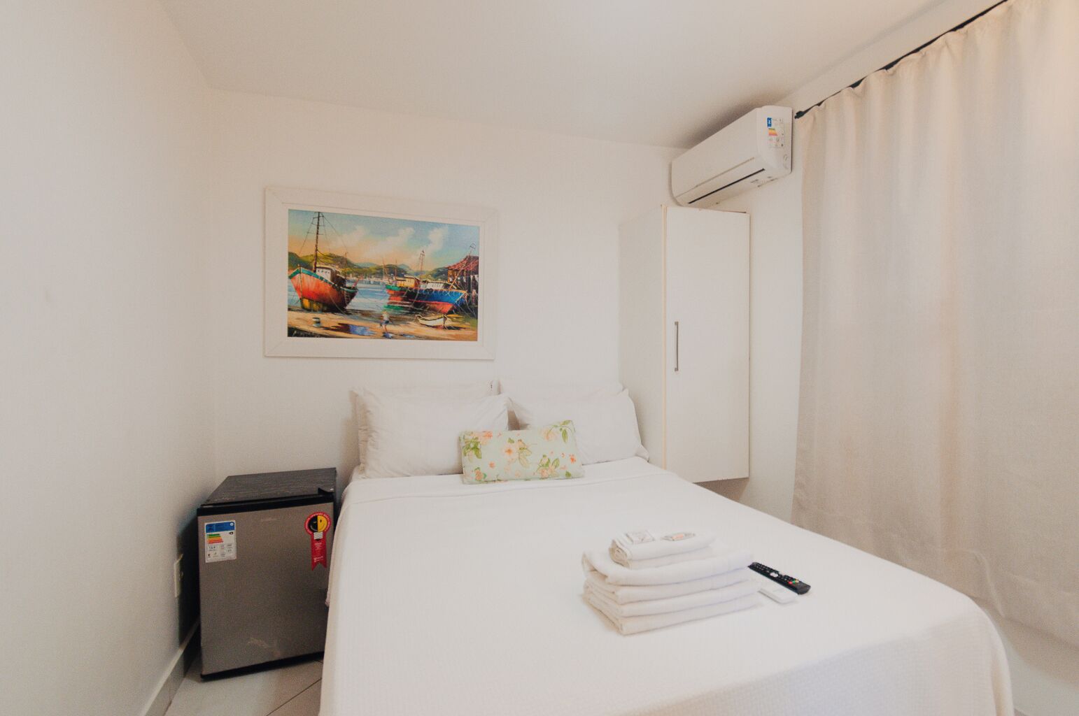 Une chambre d’hôtel avec un lit, un tableau représentant des bateaux, une armoire, un appareil de climatisation et un petit réfrigérateur.