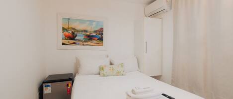A hotel room with a bed, a painting of boats, a wardrobe, an air conditioning unit, and a small refrigerator.