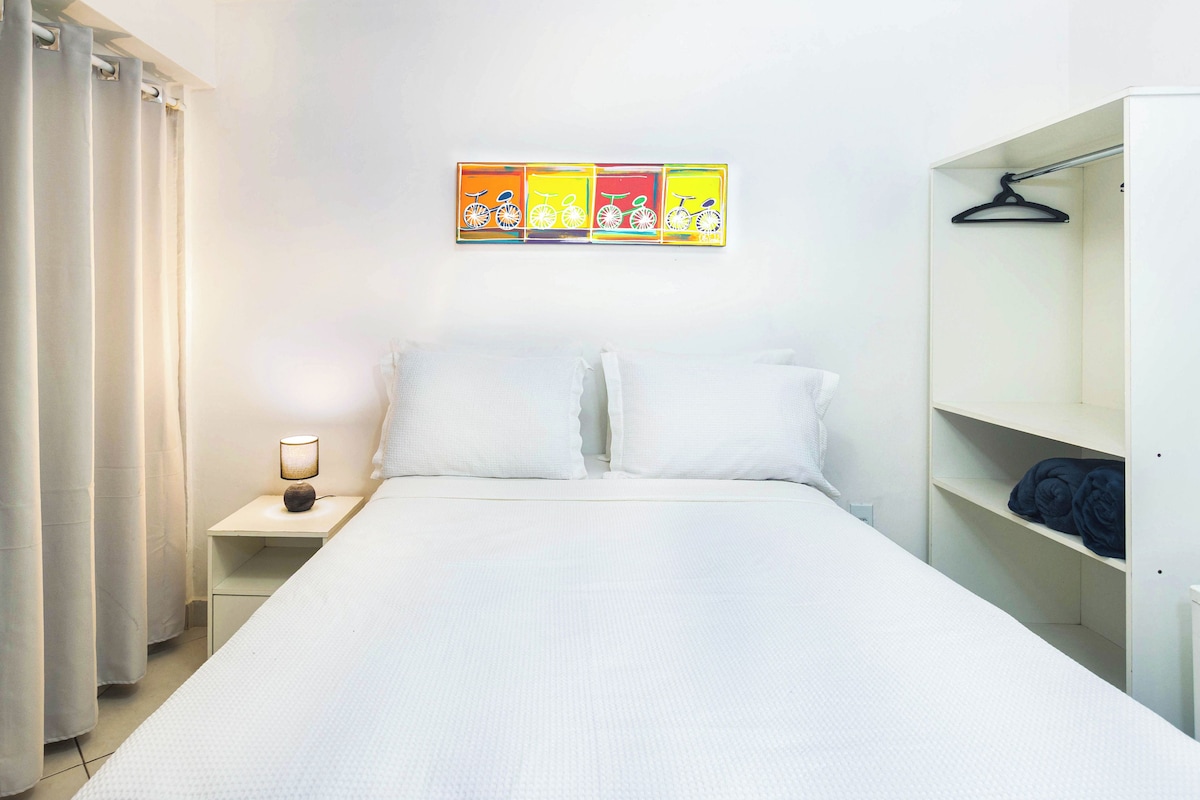 A hotel room with a bed, a nightstand with a lamp, a wardrobe, and a wall with colorful artwork.