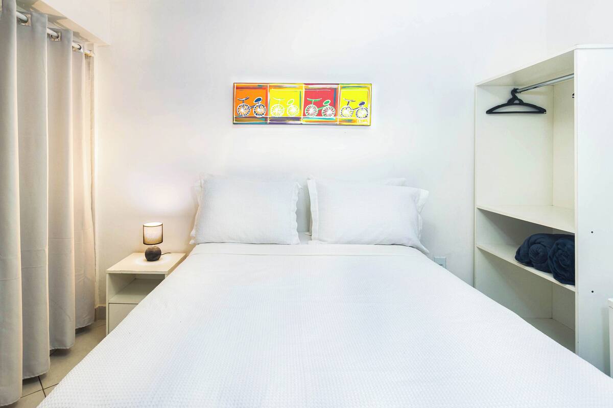 Hotellihuone, jossa on sänky, yöpöytä lampun kanssa, vaatekaappi ja seinä, jossa on värikkäitä taideteoksia.