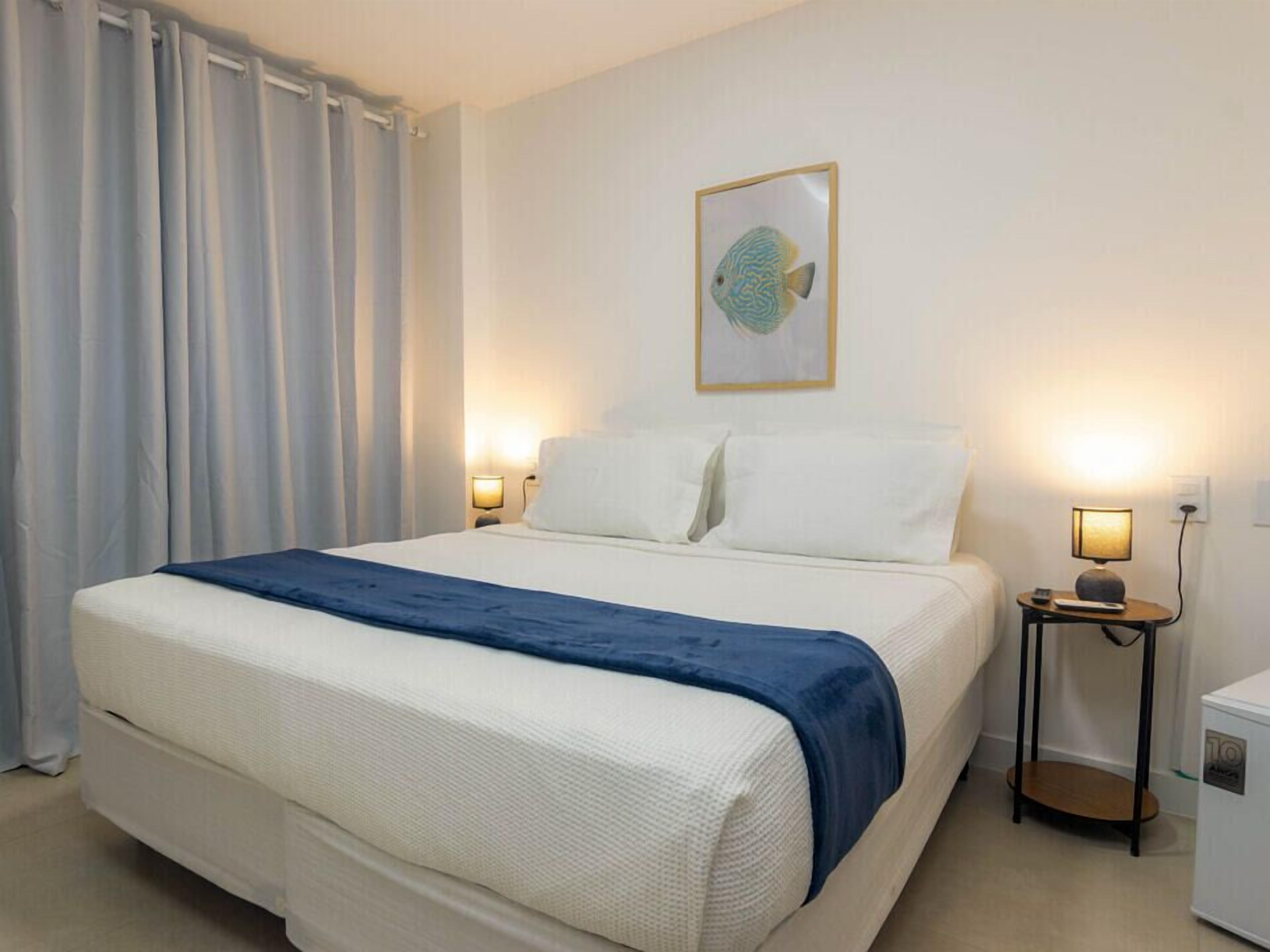 Habitación de hotel con cama, dos lámparas de noche, una obra de arte en la pared y una mesita con lámpara.