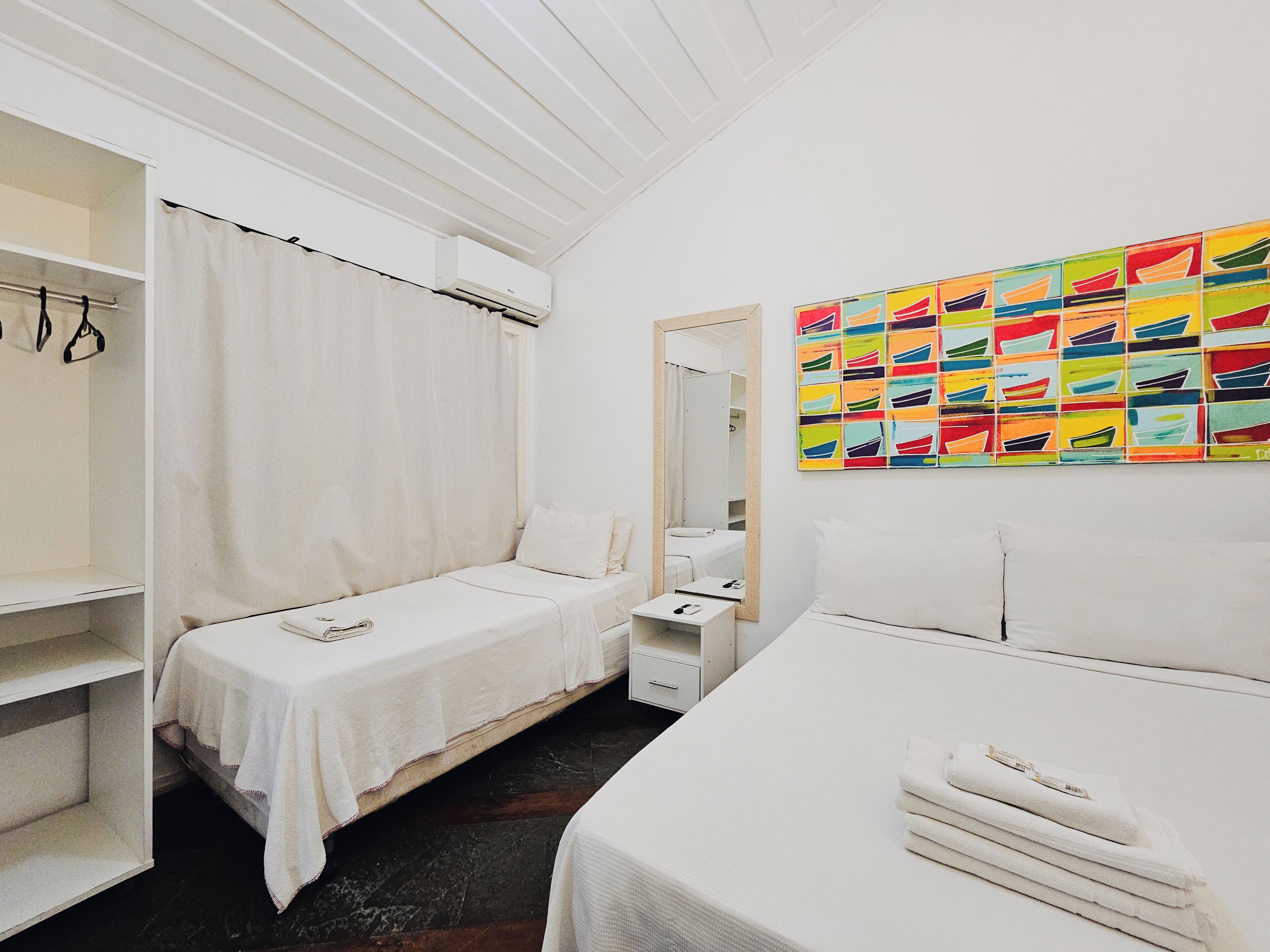 Ein Hotelzimmer mit zwei Einzelbetten, einem Schrank, einem kleinen Tisch mit Spiegel und einem farbenfrohen Bild an der Wand.