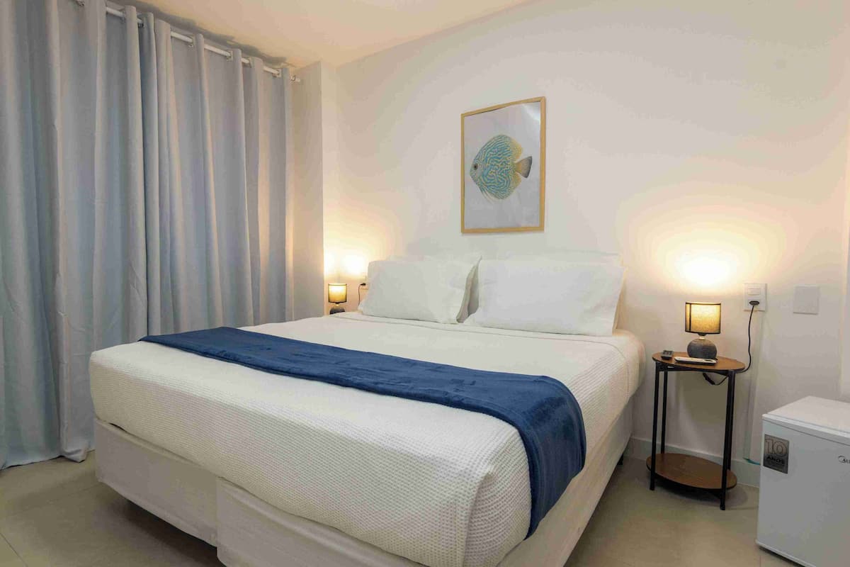 A hotel room with a bed, two bedside lamps, a small table, a nightstand, and a wall-mounted picture.