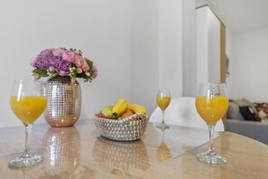 Una mesa con dos vasos de jugo de naranja, un florero con flores rosadas y una cesta de fruta.