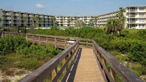 A wooden boardwalk leading to a beachfront hotel with multiple balconies and a pool area.