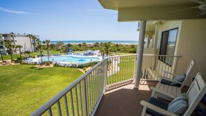A balcony with a view of a pool and beach, white railings, and two rocking chairs.