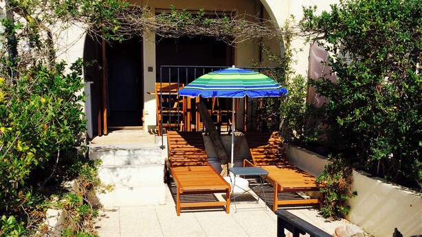 An outdoor seating area with wooden tables and chairs, a blue umbrella, and a stone pathway leading to an entrance.