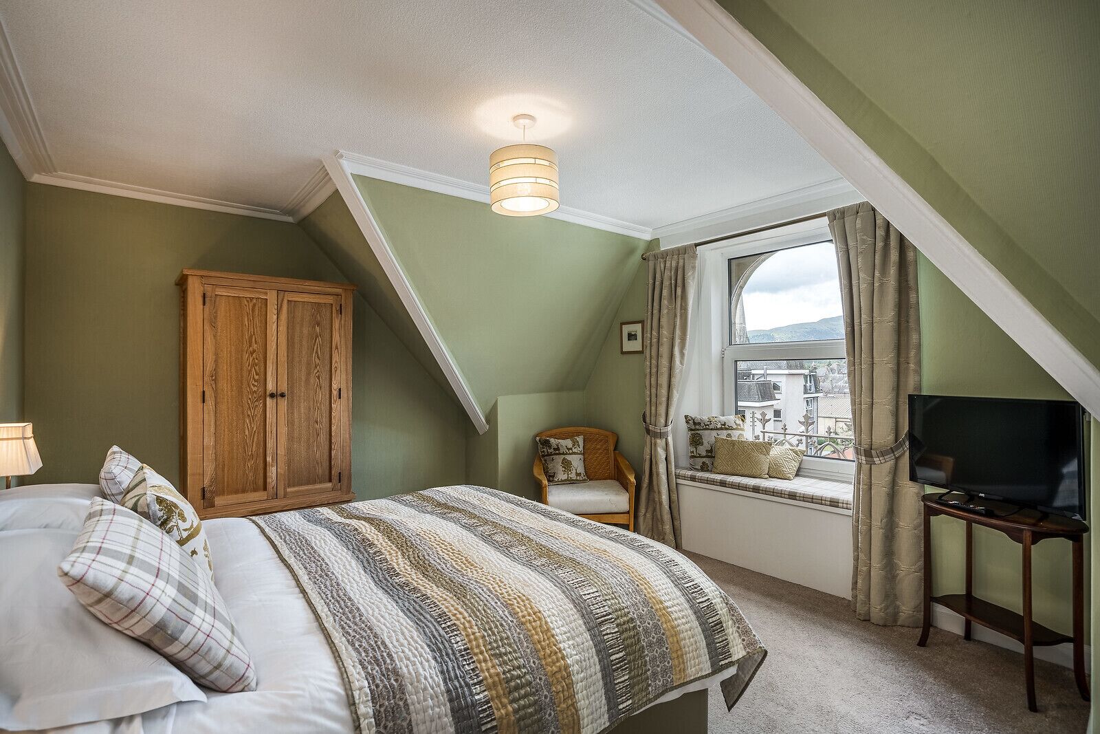 Une chambre à coucher avec un lit, une armoire en bois, un téléviseur posé sur un meuble et une fenêtre avec des rideaux.