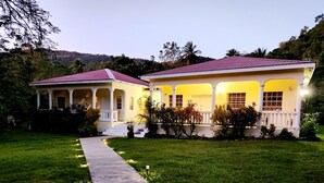 A two-story house with a red roof, white columns, and a paved pathway leading to the entrance. - Maison 79 – Your Private Soufrière Escape w/ 2BR, 2BA, AC & WiFi (Soufrière)