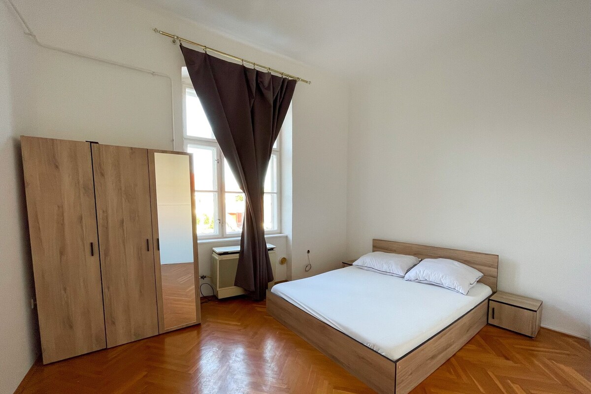 A bedroom with a wooden floor, a bed with white bedding, a wardrobe, and a window with curtains.