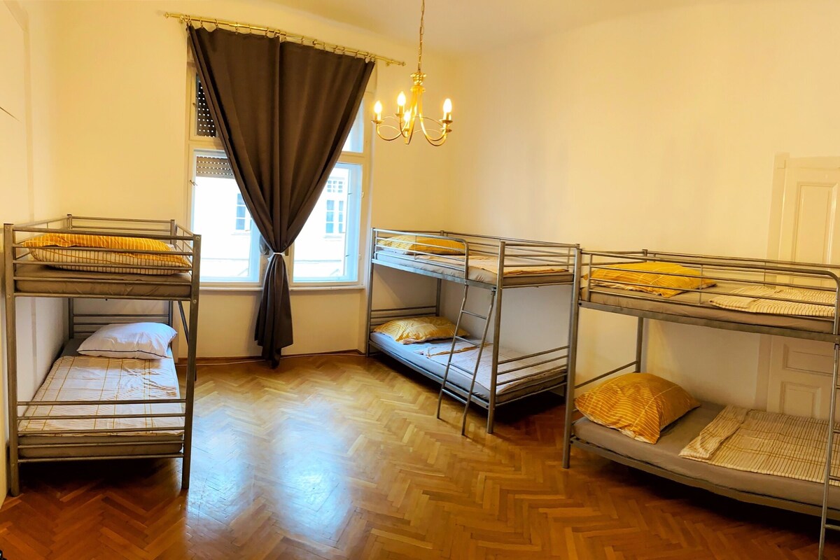 A dormitory room with bunk beds, a window with curtains, a chandelier, and a wooden parquet floor.