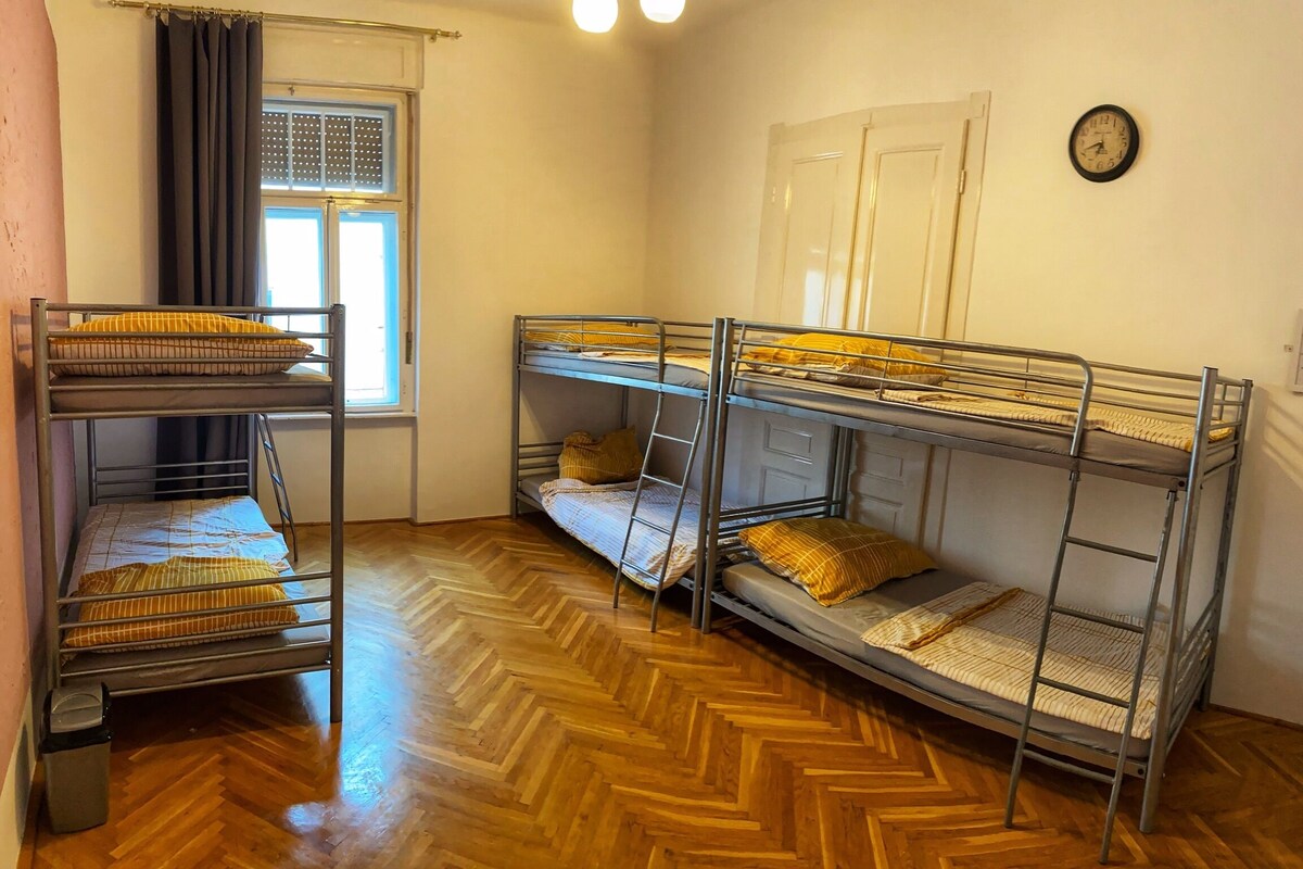A dormitory room with bunk beds, a window with curtains, a clock on the wall, and a wooden herringbone floor.