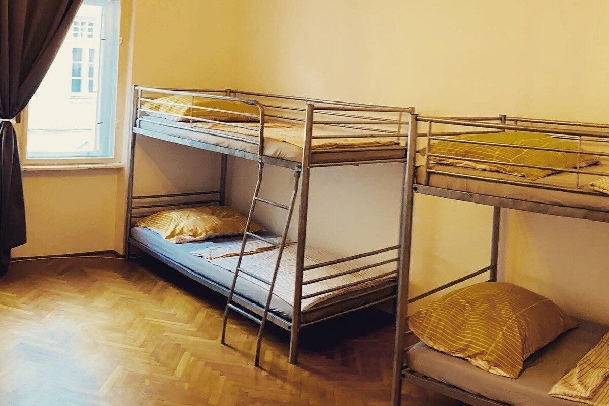 A room with two bunk beds, a wooden herringbone floor, a window with curtains, and a chandelier.
