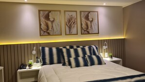 A modern bedroom with a large bed, striped pillows, a bedside table with a lamp, and three framed wall art pieces. - GOLDFLAT CABO BRANCO JOÃO PESSOA (João Pessoa)