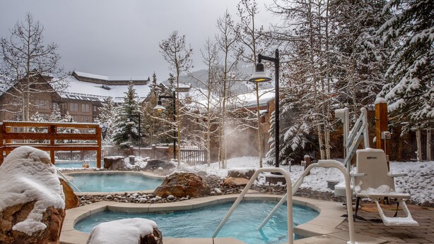 A snow-covered outdoor pool area with a hot tub, lounge chairs, and a wooden deck.