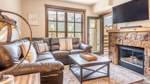 A modern living room with a leather sofa, a fireplace, a television, and a large window with a mountain view.