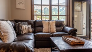 A living room with a dark leather sofa, a wooden coffee table, and a large window with a mountain view.
