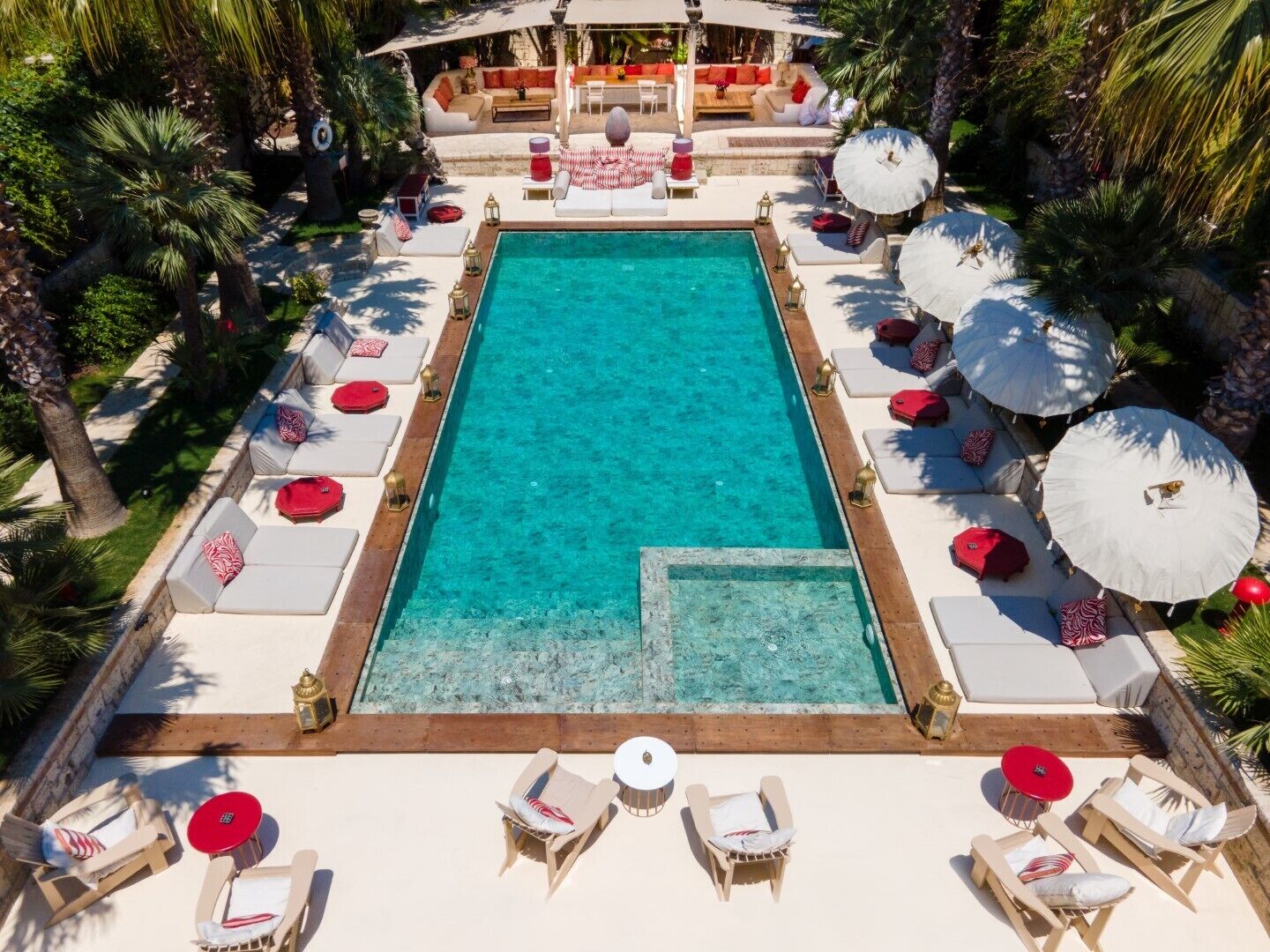 A rectangular swimming pool with clear blue water, surrounded by lounge chairs and umbrellas, and a seating area with sofas and a table in the background.
