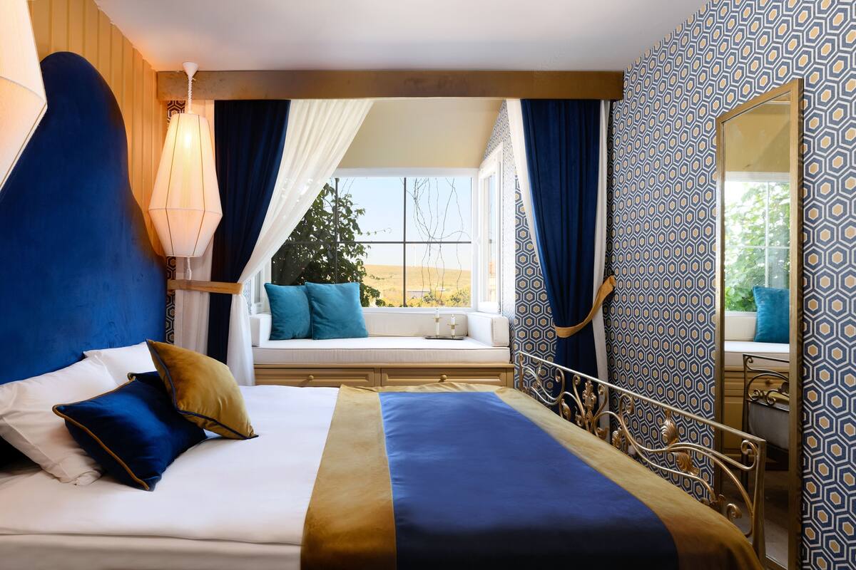 A bedroom with a large bed, a window seat, a mirror, and patterned wallpaper.