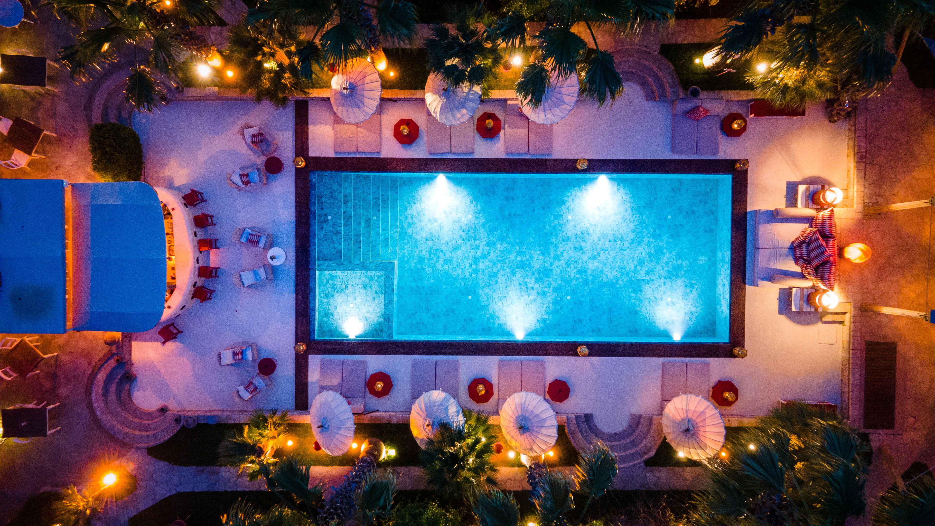 Une piscine rectangulaire agrémentée d’une fontaine, entourée de chaises longues et de parasols, avec des palmiers et un éclairage extérieur.