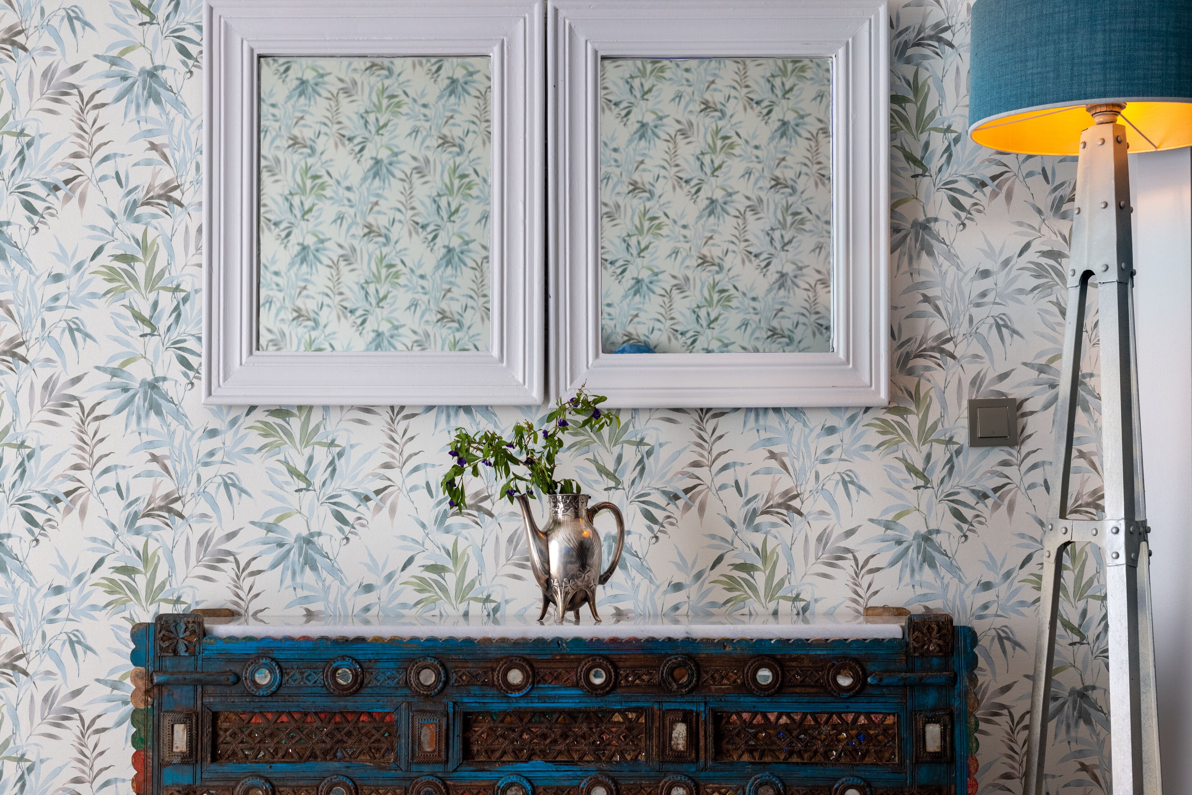 Una habitación con papel tapiz estampado, un jarrón decorativo con una planta y un espejo con marco azul.