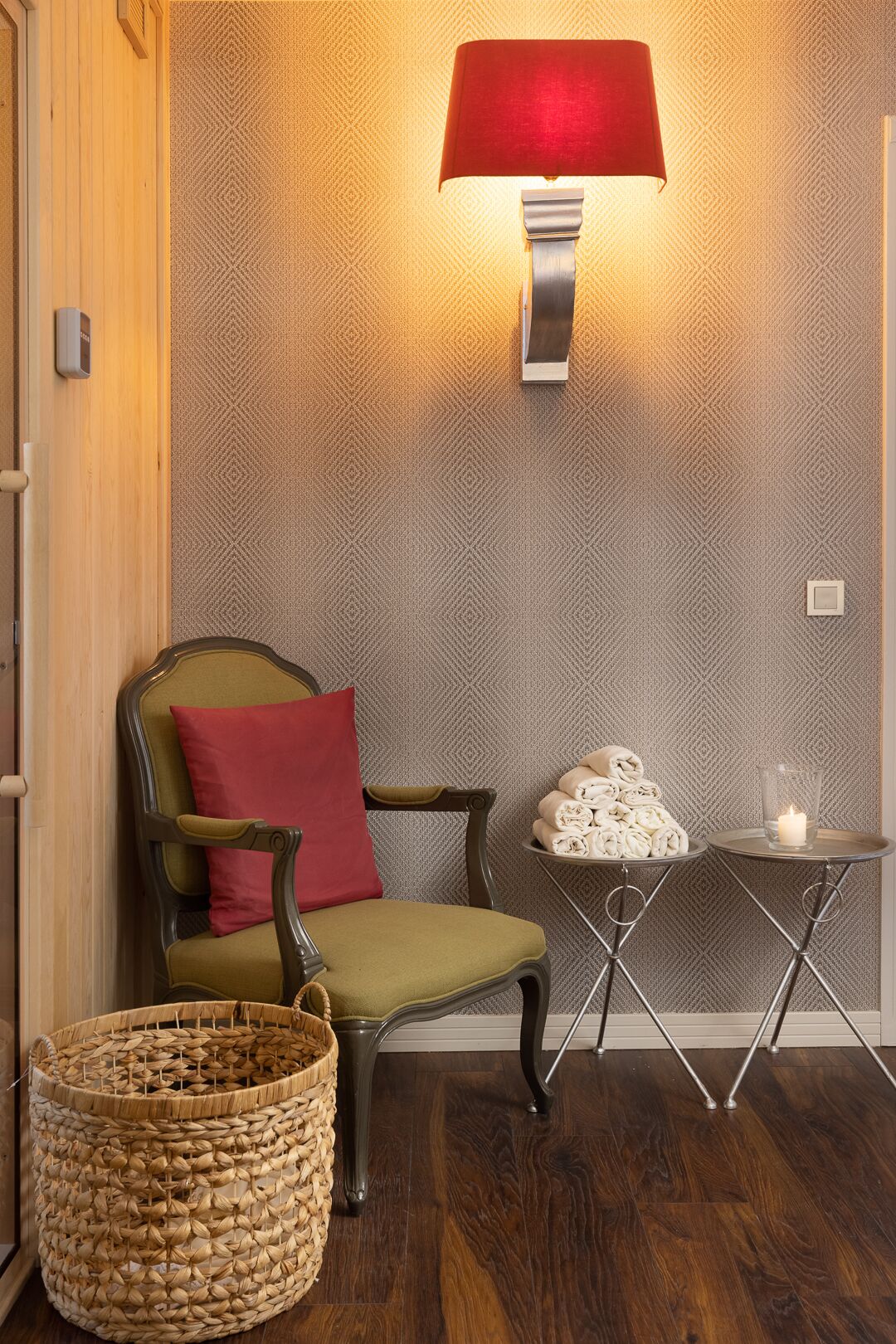 A cozy room with a patterned wall, a green armchair with a red pillow, a small table with a candle, and a woven basket.