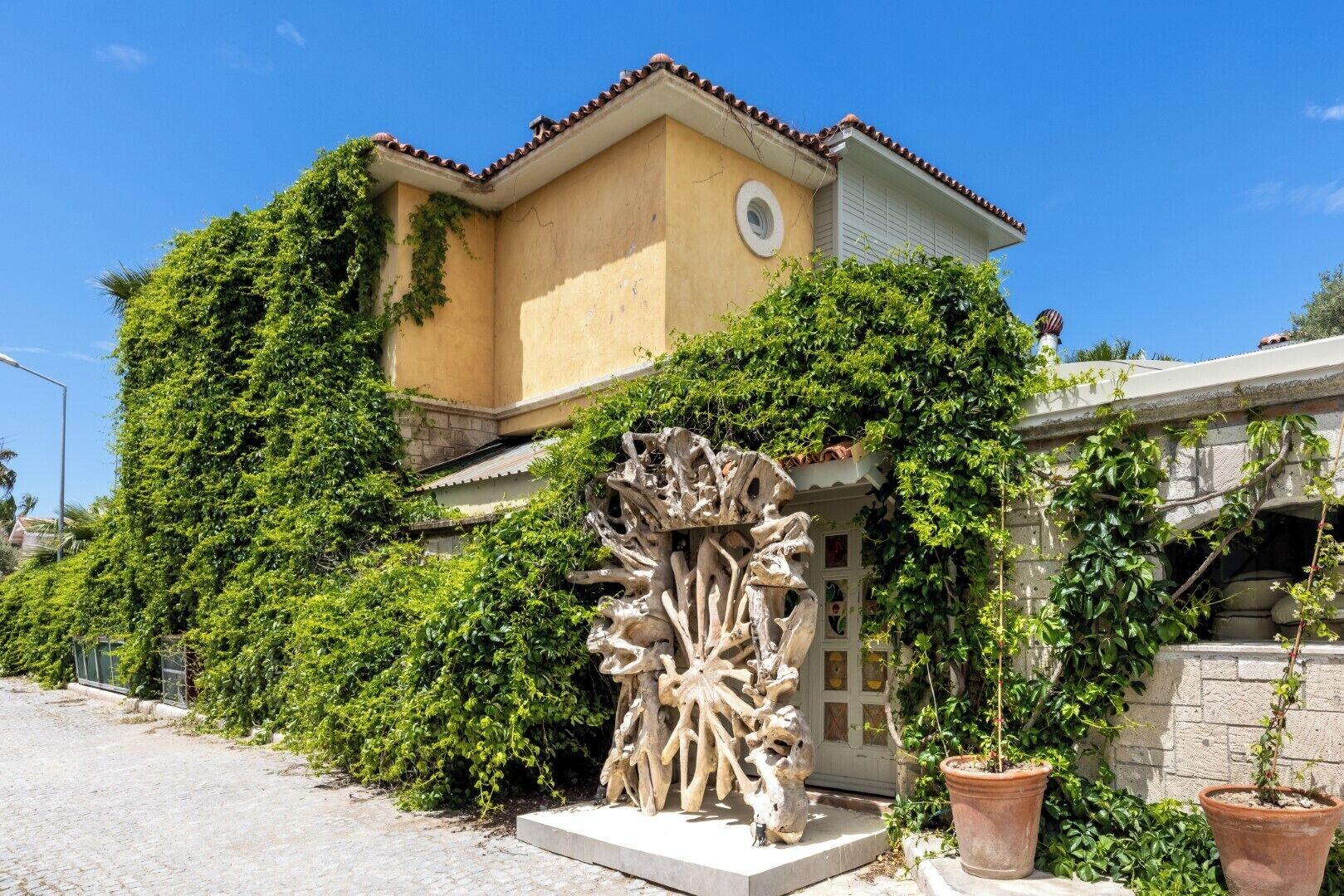 A building with a yellow facade, a red-tiled roof, and a large tree-like sculpture in front.