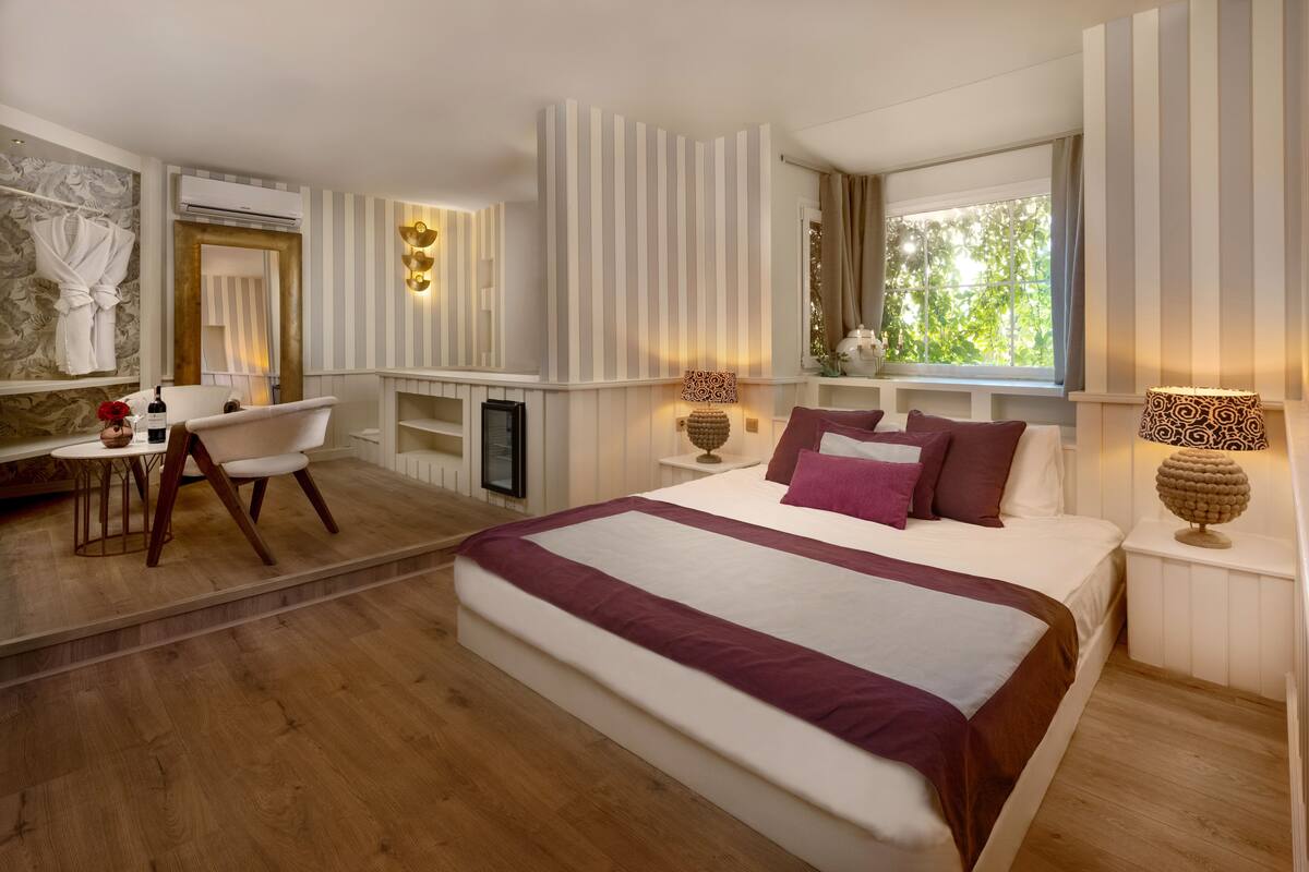 A modern hotel room with a large bed, a sitting area with a chair, a small table, and a window with a view of greenery.